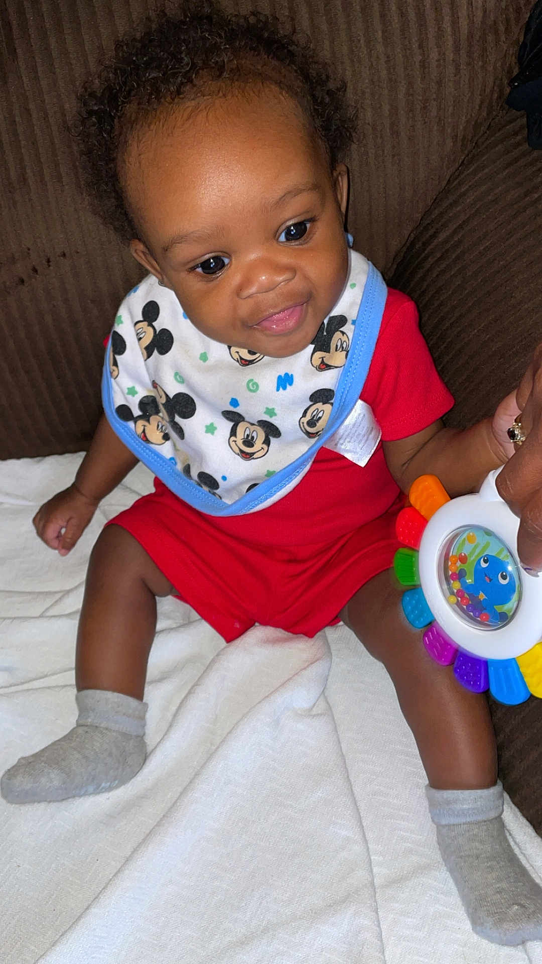 Andreus is registered to the contest to win money with this photo: baby, child, smiling, red_clothing, bib, mickey_mouse, toy, colorful, sitting, blanket, couch, happy, indoor, sock, person, cute, infant, playful, hand, face
