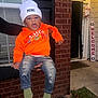 beanie, brick_wall, casual, child, clothing, cute, door, expression, green_socks, hand, holding, orange_sweatshirt, outdoor, person, porch, ripped_jeans, toddler, welcome_sign, white_hat, window