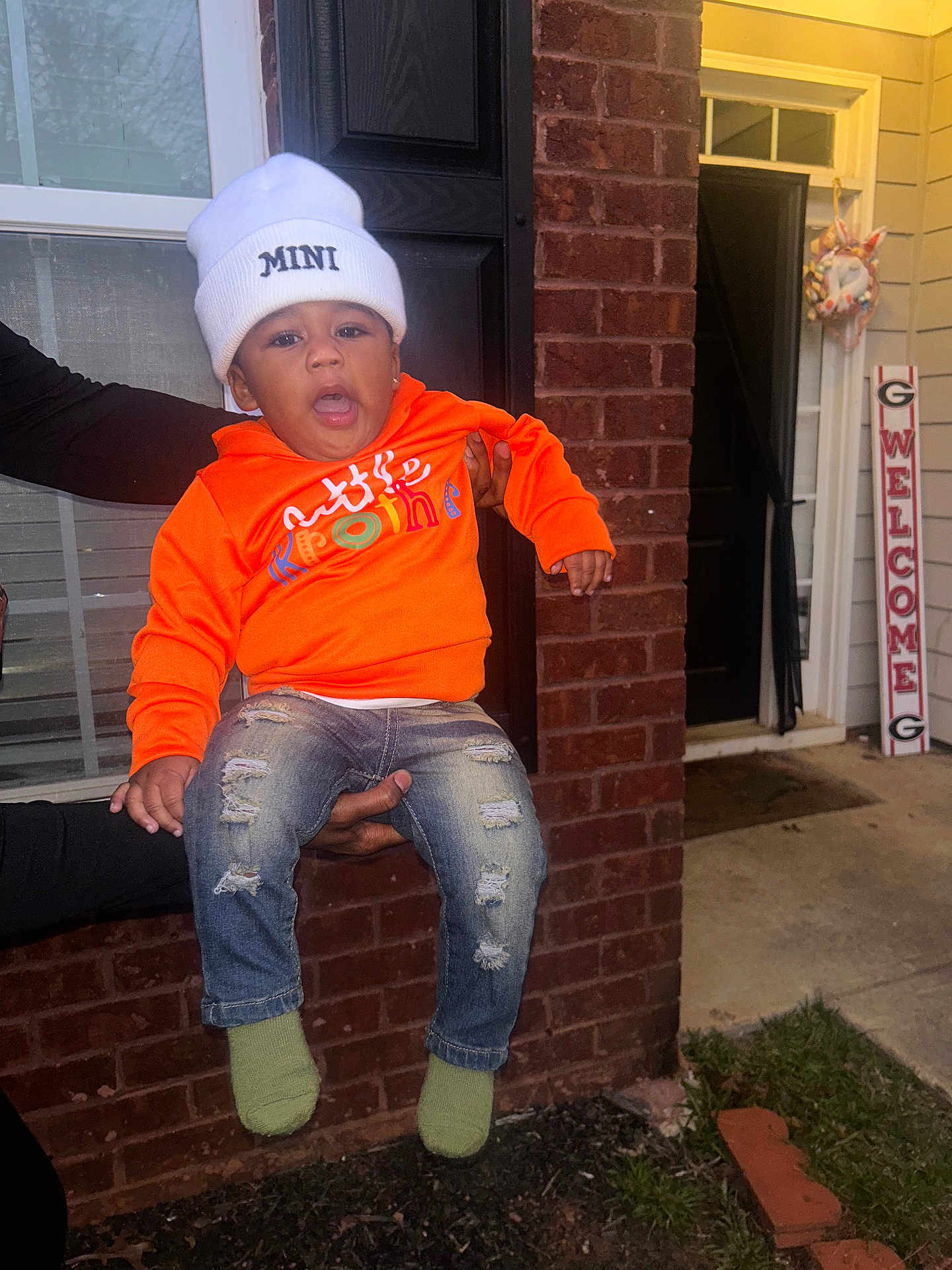 Lamajai is registered to the contest to win money with this photo: beanie, brick_wall, casual, child, clothing, cute, door, expression, green_socks, hand, holding, orange_sweatshirt, outdoor, person, porch, ripped_jeans, toddler, welcome_sign, white_hat, window