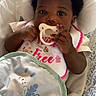 baby, infant, pacifier, bib, headband, cute, patterned_baby_seat, hand, face, eyes, clothing, soft_lighting, indoor, child, adorable, newborn, portrait, cozy, resting, young