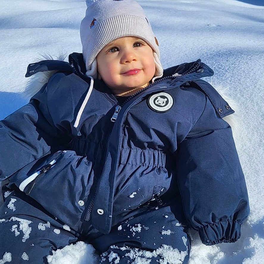Jahyah a rejoint le concours — aidez-le/la à gagner de superbes lots ! baby, beanie, boots, child, cold_weather, hat, houses, outdoor, play, portrait, rosy_cheeks, sitting, smile, snow, snowsuit, snowy_landscape, sunlight, toddler, trees, winter_coat
