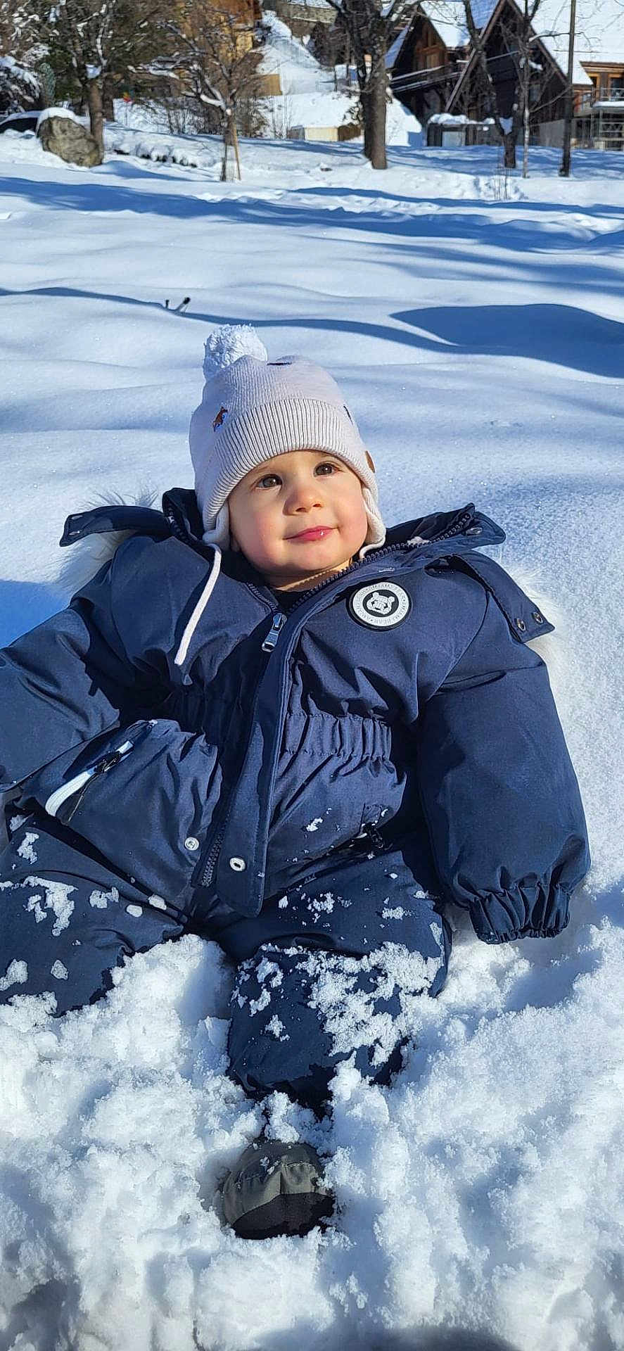 Jahyah a rejoint le concours — aidez-le/la à gagner de superbes lots ! baby, beanie, boots, child, cold_weather, hat, houses, outdoor, play, portrait, rosy_cheeks, sitting, smile, snow, snowsuit, snowy_landscape, sunlight, toddler, trees, winter_coat