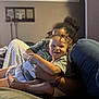 Giovanni is registered to the contest to win money with this photo: toddler, child, smiling, curly_hair, sitting, adult, couch, blanket, indoor, happy, holding_object, person, home, relaxing, cozy, furniture, casual_clothing, portrait, family, living_room