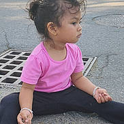 Sanu is registered to the contest to win money with this photo: child, girl, pink_shirt, black_pants, sitting, outdoor, street, drain, bracelet, casual_clothing, curly_hair, side_view, footwear, asphalt, gravel, nature_background, young_child, thoughtful, earring, daylight