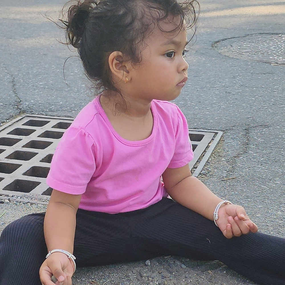 Sanu is registered to the contest to win money with this photo: asphalt, black_pants, bracelet, casual_clothing, child, curly_hair, daylight, drain, earring, footwear, girl, gravel, nature_background, outdoor, pink_shirt, side_view, sitting, street, thoughtful, young_child