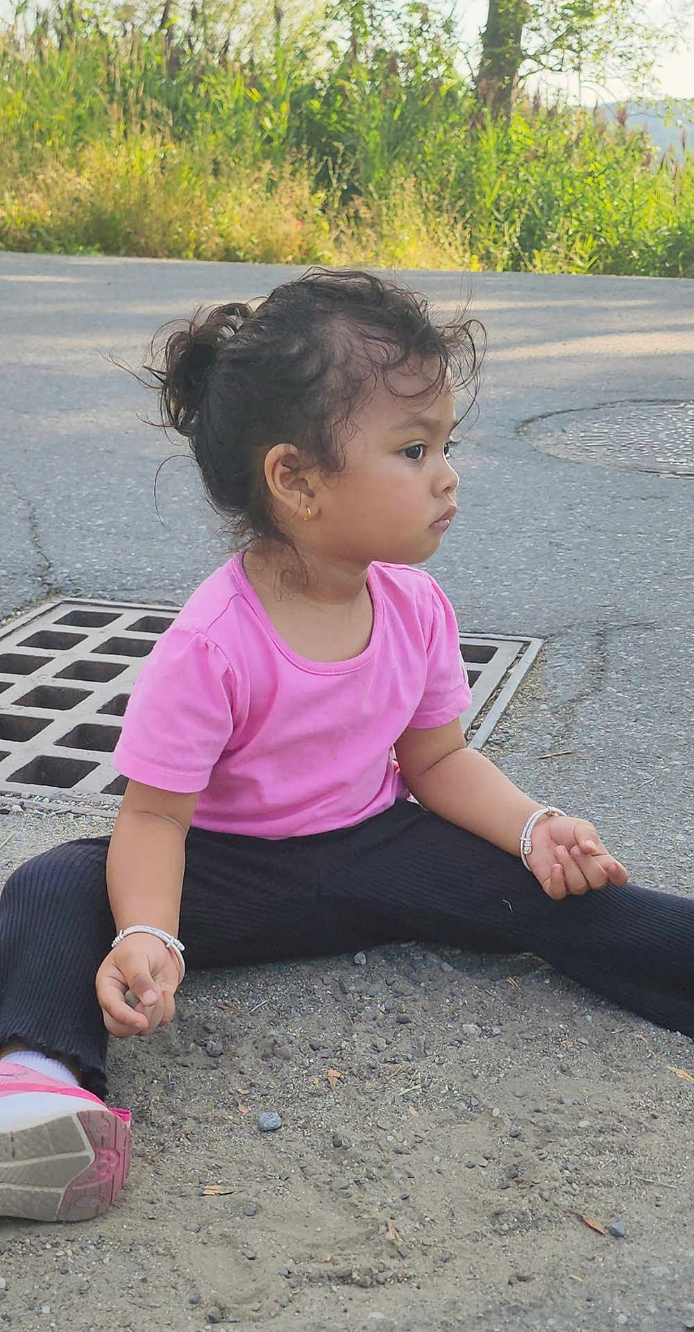 Sanu is registered to the contest to win money with this photo: child, girl, pink_shirt, black_pants, sitting, outdoor, street, drain, bracelet, casual_clothing, curly_hair, side_view, footwear, asphalt, gravel, nature_background, young_child, thoughtful, earring, daylight