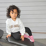 Sanu is registered to the contest to win money with this photo: bench, black_leggings, casual, child, clothing, curly_hair, fashion, footwear, gray_wall, indoor, los_angeles, person, pink_sneakers, portrait, resting, serious_expression, toddler, white_sweatshirt, workout_bench, young_child