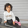 bench, black_leggings, casual, child, clothing, curly_hair, fashion, footwear, gray_wall, indoor, los_angeles, person, pink_sneakers, portrait, resting, serious_expression, toddler, white_sweatshirt, workout_bench, young_child