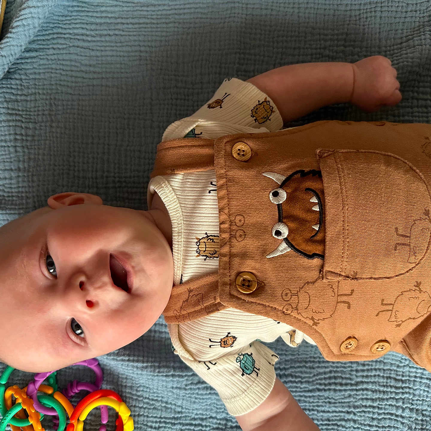 Rune joined the competition — help win amazing prizes! baby, infant, smiling, lying_down, overalls, monster_design, shirt, blue_blanket, toy, colorful, plastic_rings, cute, child, happy, indoors, clothing, portrait, one_person, casual, playful