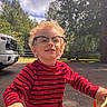 child, boy, glasses, curly_hair, red_shirt, striped_shirt, tricycle, outdoor, sunny, trees, vehicle, smiling, play, daytime, parked_car, nature, happiness, person, young, recreation