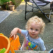 Howard is registered to the contest to win money with this photo: toddler, child, curly_hair, outdoor, toy, orange, container, potted_plant, folding_chair, concrete, play, smile, casual_clothing, young_child, sunny_day, garden, person, happy, blue_eyes, short_sleeves