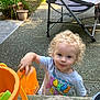 toddler, child, curly_hair, outdoor, toy, orange, container, potted_plant, folding_chair, concrete, play, smile, casual_clothing, young_child, sunny_day, garden, person, happy, blue_eyes, short_sleeves