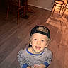 child, boy, curly_hair, cap, striped_pajamas, wooden_floor, table, chairs, indoor, smiling, holding_object, looking_up, blond, young_child, casual_clothing, home_interior, shadow, warm_lighting, person, happy