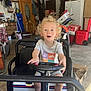 child, toddler, toy_car, steering_wheel, garage, curly_hair, excited, open_mouth, white_shirt, shorts, floor, boxes, refrigerator, chair, door, table, storage, red_container, window, person