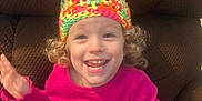 Taryn is registered to the contest to win money with this photo: child, toddler, smiling, curly_hair, knitted_hat, colorful_hat, pink_sweater, happy, waving, indoor, chair, brown_chair, cute, person, portrait, seated, joyful, young_child, casual_clothing, bright_colors