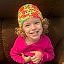 Taryn joined the competition — help win amazing prizes! toddler, child, smiling, curly_hair, colorful_hat, pink_sweater, couch, indoor, happy, portrait, cute, young_child, seated, cozy, casual_clothing, face, person, warm_lighting, playful, expressive