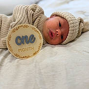 Walker is registered to the contest to win money with this photo: baby, newborn, infant, knitted_clothing, hat, plaque, milestone, one_month, blanket, lying_down, soft_lighting, beige, cream, portrait, cute, face, headwear, cozy, closeup, resting