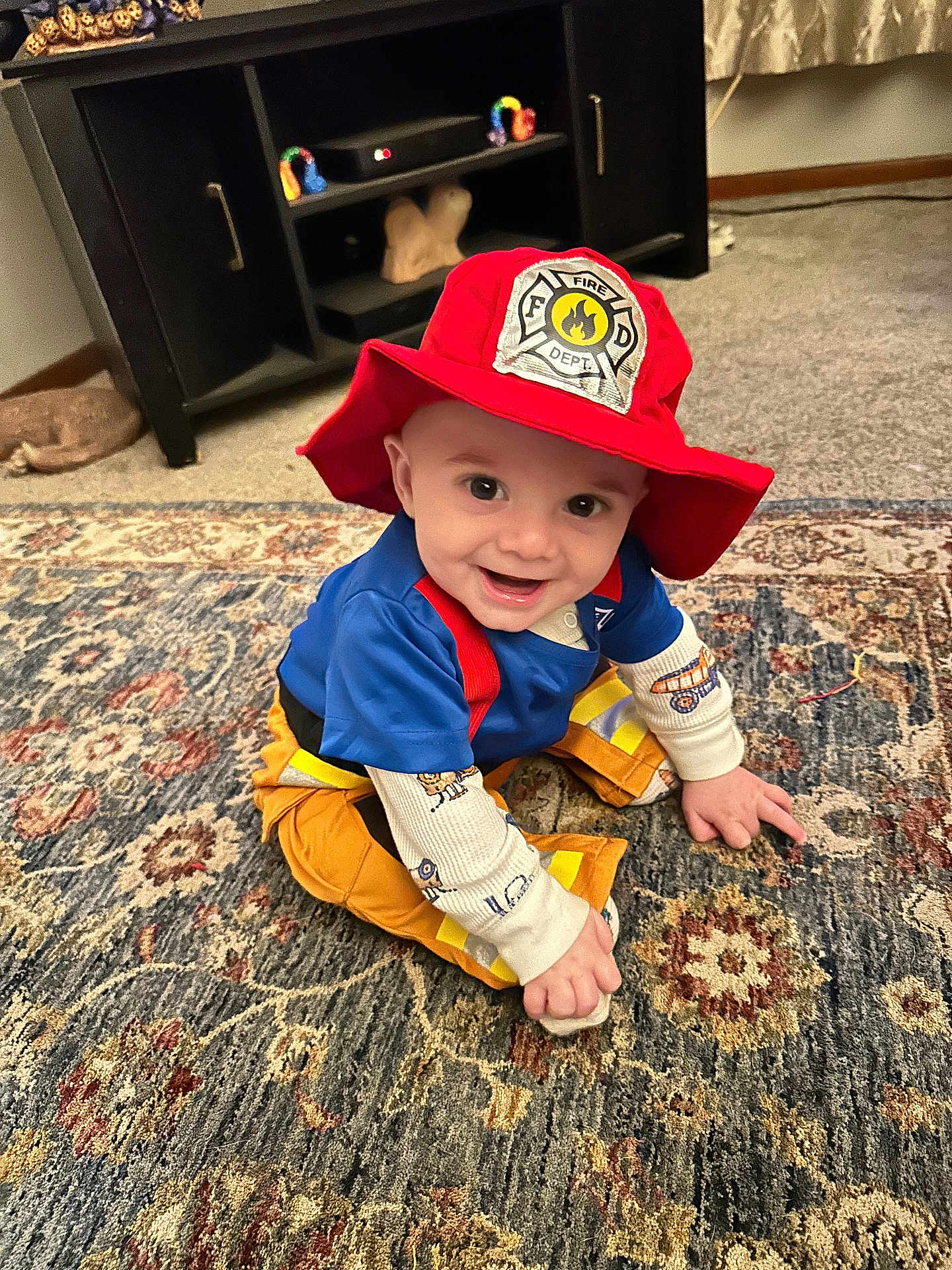 Cooper joined the competition — help win amazing prizes! baby, child, smiling, firefighter_hat, costume, indoor, rug, carpet, blue_shirt, orange_pants, long_sleeves, person, cute, happy, sitting, floor, home, decor, toy, face
