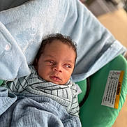 Josiah joined the competition — help win amazing prizes! newborn, baby, infant, child, sleepy, side_eye, face, portrait, blanket, swaddle, clothing, green_seat, baby_carrier, indoors, soft_lighting, curly_hair, brown_skin, eyes, cozy, warning_label