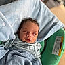 newborn, baby, infant, child, sleepy, side_eye, face, portrait, blanket, swaddle, clothing, green_seat, baby_carrier, indoors, soft_lighting, curly_hair, brown_skin, eyes, cozy, warning_label