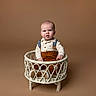 baby, brown_background, child, cute, expression, fashion, footwear, furniture, indoor, infant, neutral_expression, one_person, overalls, person, portrait, shirt, sitting, studio_photo, wicker_basket, young_child