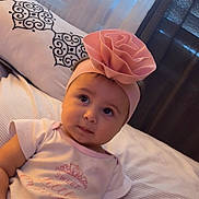 Eliana is registered to the contest to win money with this photo: baby, child, headband, pink, flower, bed, pillow, pattern, curious, infant, clothing, toddler, indoor, portrait, cute, soft_light, person, sleepwear, smile, young