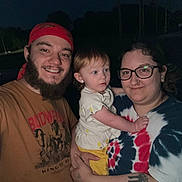 Ariah is registered to the contest to win money with this photo: smiling, beard, bandana, glasses, toddler, child, family, outdoor, night, flash, person, portrait, casual_clothing, tattoo, holding, happy, tshirt, tie_dye, headwear, light_skin