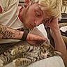 affection, animal, bed, blonde_hair, casual_clothing, cat, close_up, cozy, ear_piercing, indoor, kitten, lying_down, person, pet, relaxed, striped_cat, tattoo, tattooed_arm, watch, young_adult