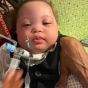 Jahlil is registered to the contest to win money with this photo: baby, child, medical_equipment, breathing_tube, vest, shirt, hand, face, tongue, closeup, person, indoor, patterned_background, adult_hand, medical_care, infant, cute, healthcare, expression, support