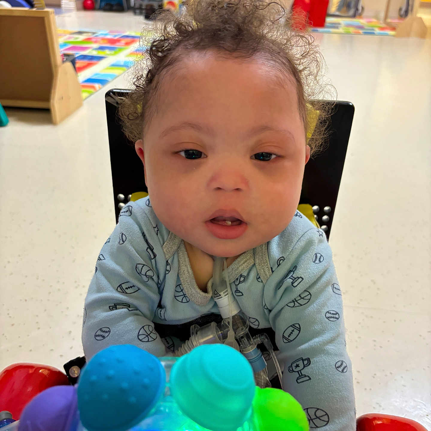 Jahlil joined the competition — help win amazing prizes! baby, blue_onesie, blurred_background, child, colorful, curly_hair, cute, floor, flooring, healthcare_tube, home, indoor, infant, medical_device, person, plastic_toy, play_area, red_chair, toy, woman