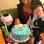 birthday_cake, blue_cake, sprinkles, toddler, child, woman, helmet, toy, breathing_tube, phone, hand, couch, celebration, party, indoor, person, holding, sitting, happy_birthday, colorful