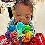 baby, child, toy, colorful, curly_hair, indoor, playroom, focus, light, plastic, hands, face, sitting, table, red, blue, green, yellow, person, infant