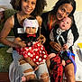 two_women, two_babies, baby_helmet, polka_dot_dress, medical_equipment, hospital_room, smiling_women, seated, holding_babies, curly_hair, ripped_jeans, red_dress, black_top, medical_tubes, indoor, family, caregiving, happy, infant, mother