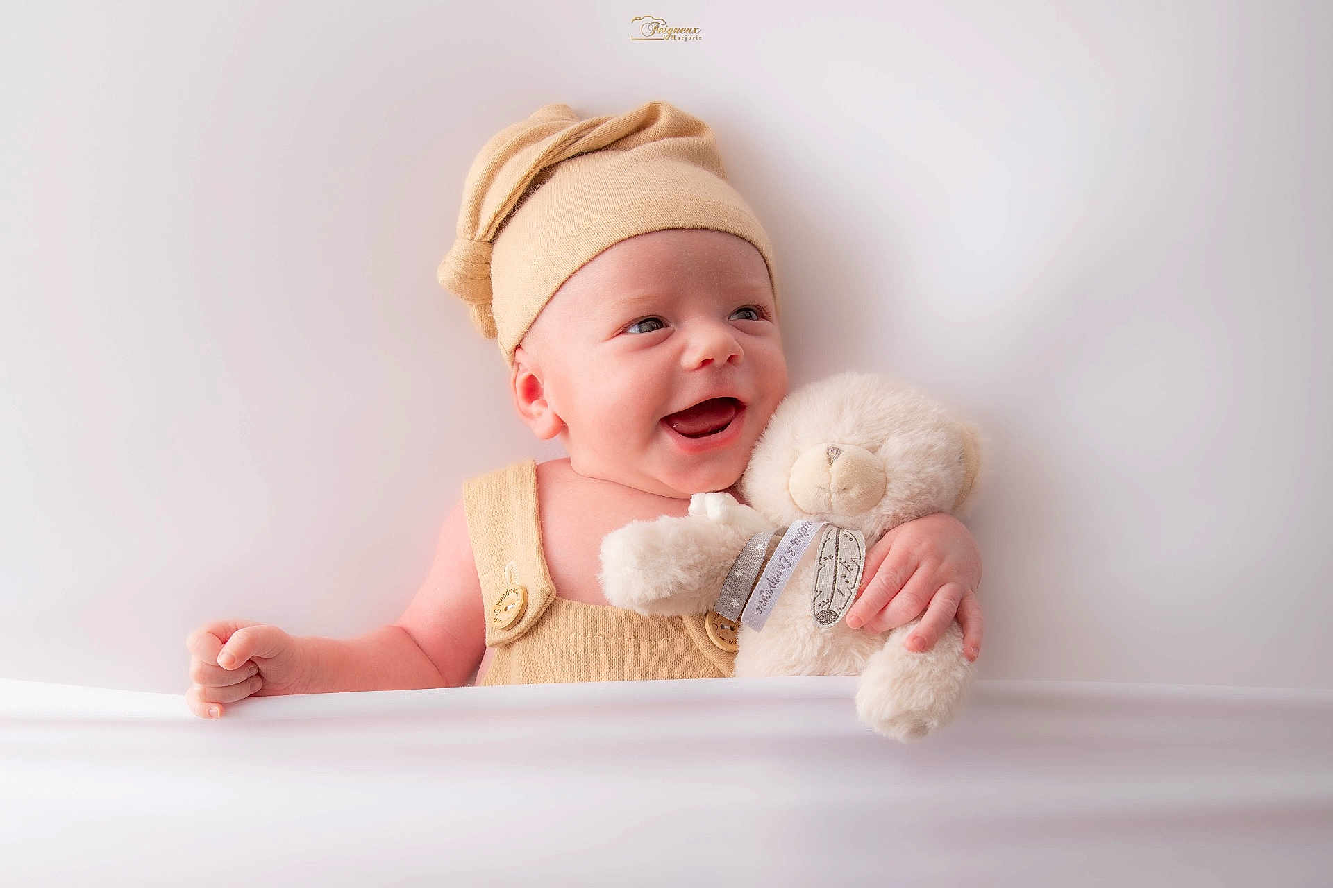Elio a rejoint le concours — aidez-le/la à gagner de superbes lots ! baby, smiling, teddy_bear, hat, beige, cute, infant, portrait, child, soft_toy, happy, cuddling, indoors, light_background, small_hand, button, fabric, warm_clothing, closeup, adorable