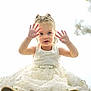 blonde_hair, blue_eyes, braids, child, cute, dress, floral_background, flower, hands_up, innocent, playful, portrait, shoes, sitting, smiling, soft_light, studio_photo, toddler, white_dress, young_child