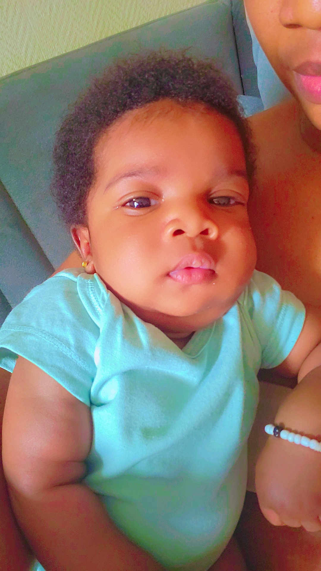 Saran participe au concours pour gagner de l'argent avec cette photo : baby, child, infant, curly_hair, blue_onesie, earring, couch, arm, adult, skin, face, portrait, closeup, holding, indoor, expressionless, person, young_child, seated, casual
