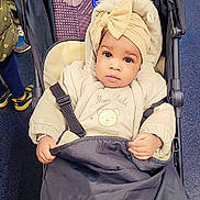 Saran Ballo a rejoint le concours — aidez-le/la à gagner de superbes lots ! baby, stroller, child, clothing, headwrap, bow, blanket, indoor, person, face, warm_clothing, seat, pattern, hand, footwear, floor, fabric, curious, black, beige