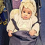 baby, beige, black, blanket, bow, child, clothing, curious, fabric, face, floor, footwear, hand, headwrap, indoor, pattern, person, seat, stroller, warm_clothing