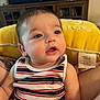baby, child, striped_clothing, tank_top, yellow_cushion, face, expression, indoor, living_room, furniture, television, person, curly_hair, skin, arm, tattoo, closeup, soft_light, candid, young