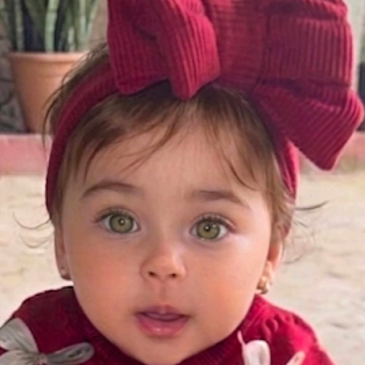 Mila participe au concours pour gagner de l'argent avec cette photo : toddler, child, face, green_eyes, red_sweater, headband, bow, cute, portrait, outdoor, plant, potted_plant, expression, curious, young_child, fashion, winter_clothing, person, baby, closeup