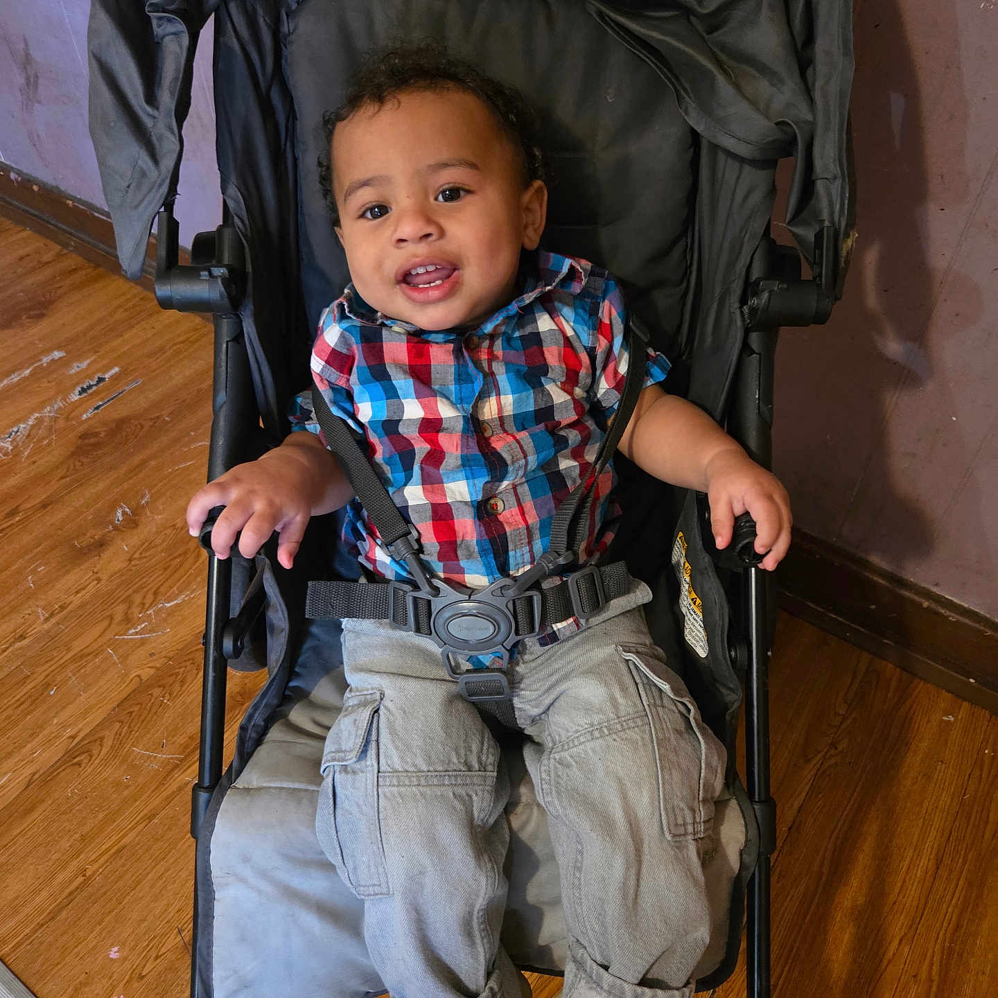 Cayden joined the competition — help win amazing prizes! toddler, child, stroller, plaid_shirt, cargo_pants, shoes, smile, indoors, wooden_floor, wall, seatbelt, black_stroller, happy, person, young_child, casual_clothing, cute, baby, sitting, portrait