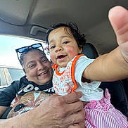 Isla Anani joined the competition — help win amazing prizes! baby, adult, smiling, car_interior, child, holding, happy, portrait, person, clothing, seat, seatbelt_warning, glasses, hand, pink_skirt, white_shirt, orange_bib, window, sunlight, closeup