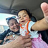 baby, adult, smiling, car_interior, child, holding, happy, portrait, person, clothing, seat, seatbelt_warning, glasses, hand, pink_skirt, white_shirt, orange_bib, window, sunlight, closeup