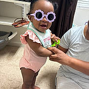 Danielle is registered to the contest to win money with this photo: toddler, child, sunglasses, indoor, carpet, person, adult_hand, curtain, window, shelf, shoes, pink_clothing, smile, standing, baby, holding_hands, toy, hair_buns, glasses, casual