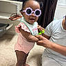 toddler, child, sunglasses, indoor, carpet, person, adult_hand, curtain, window, shelf, shoes, pink_clothing, smile, standing, baby, holding_hands, toy, hair_buns, glasses, casual