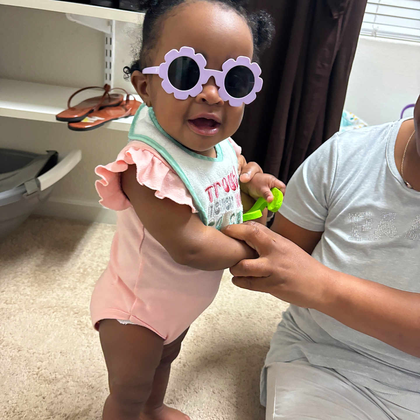 Danielle is registered to the contest to win money with this photo: toddler, child, sunglasses, indoor, carpet, person, adult_hand, curtain, window, shelf, shoes, pink_clothing, smile, standing, baby, holding_hands, toy, hair_buns, glasses, casual