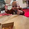 toddler, child, indoor, table, food, french_fries, box, brown_hoodie, curious, reaching, person, face, play_area, red_floor, window, signage, light, wooden_table, casual_clothing, young_child
