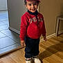 casual_clothing, child, curly_hair, door, footwear, happy, headwear, indoor, jeans, person, polo, red_cap, red_sweater, smiling, standing, tile_floor, wall, white_shoes, wooden_floor, young
