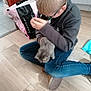 boy, kitten, cat, child, glasses, floor, wooden_floor, jeans, sitting, indoor, bag, chair, sweatshirt, pet, cute, young, home, casual, holding, animal