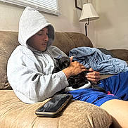 Nova is registered to the contest to win money with this photo: person, hoodie, kitten, black_cat, couch, blanket, smartphone, hand, window, blind, indoor, casual_clothing, relaxing, seated, brown_furniture, portrait, young_adult, comfort, pet, home